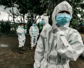 People in white protective suits and blue face masks standing on a paved area