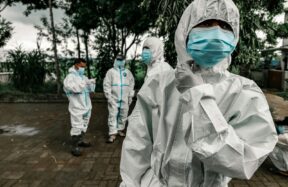 People in white protective suits and blue face masks standing on a paved area