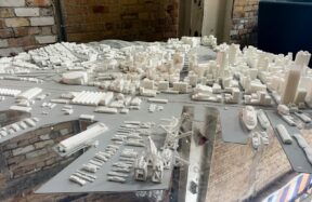 A white architectural model of a city skyline and harbor on a reflective surface