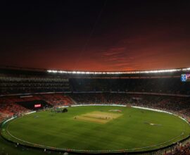 A cricket stadium with a green pitch under a dark red evening sky