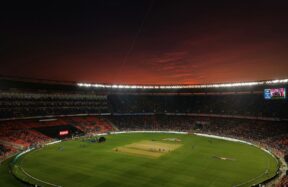 A cricket stadium with a green pitch under a dark red evening sky