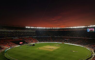 A cricket stadium with a green pitch under a dark red evening sky
