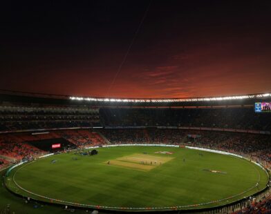 A cricket stadium with a green pitch under a dark red evening sky