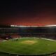 A cricket stadium with a green pitch under a dark red evening sky