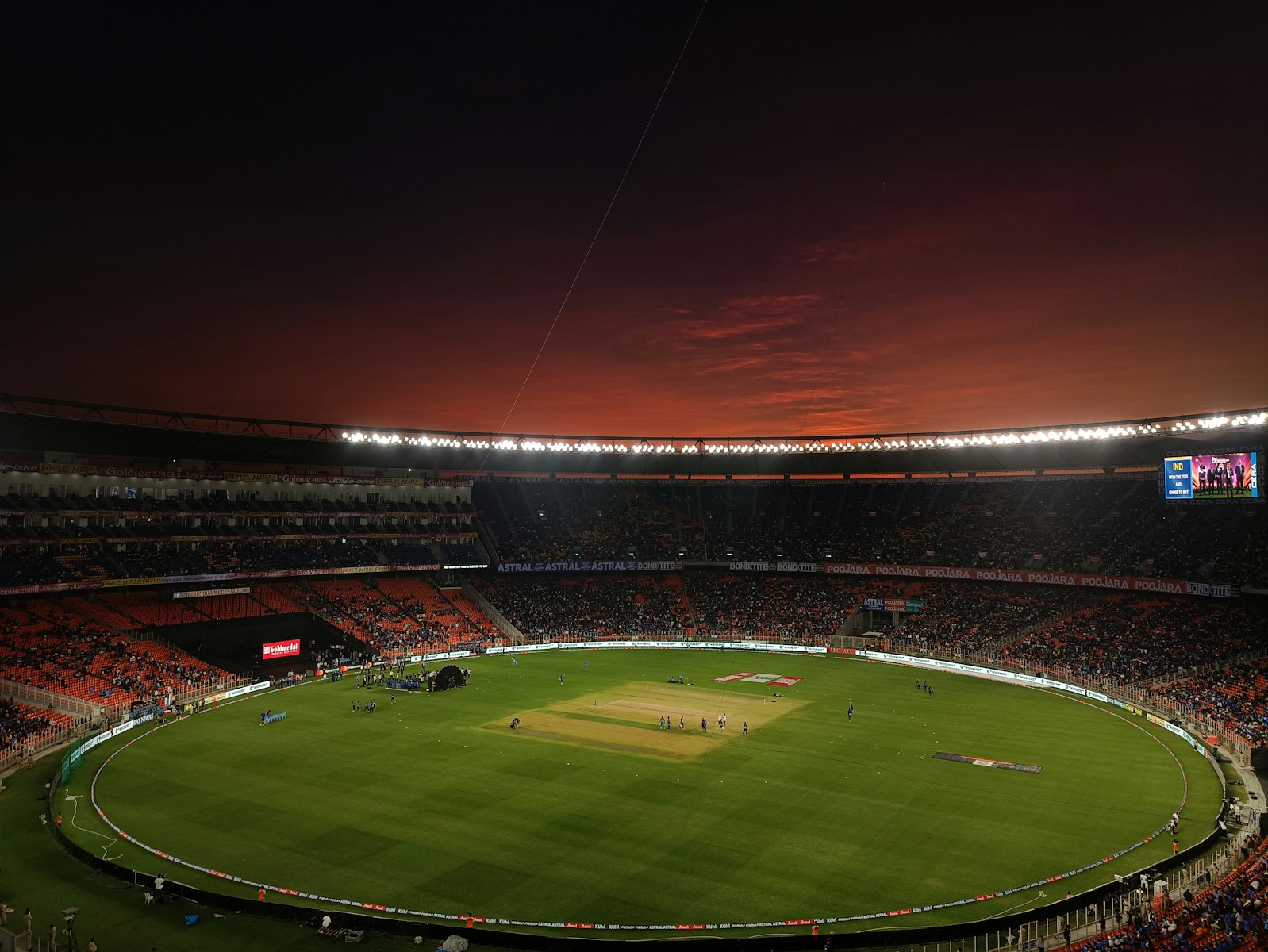 A cricket stadium with a green pitch under a dark red evening sky
