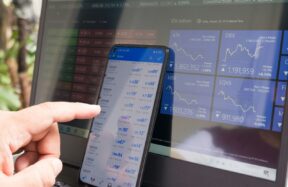 A hand pointing at a smartphone displaying currency quotes against a laptop