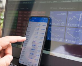 A hand pointing at a smartphone displaying currency quotes against a laptop