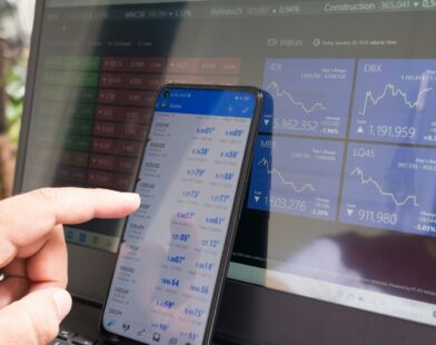 A hand pointing at a smartphone displaying currency quotes against a laptop