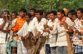 Many men standing outdoors, some holding small objects