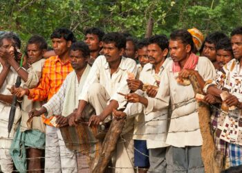 Many men standing outdoors, some holding small objects